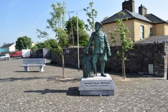 A statue of General Sean Mac Eoin stands on a stone plinth in a paved courtyard, with small trees and a yellow building in the background.