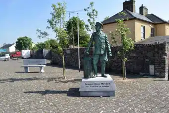 A statue of General Sean Mac Eoin stands on a stone plinth in a paved courtyard, with small trees and a yellow building in the background.