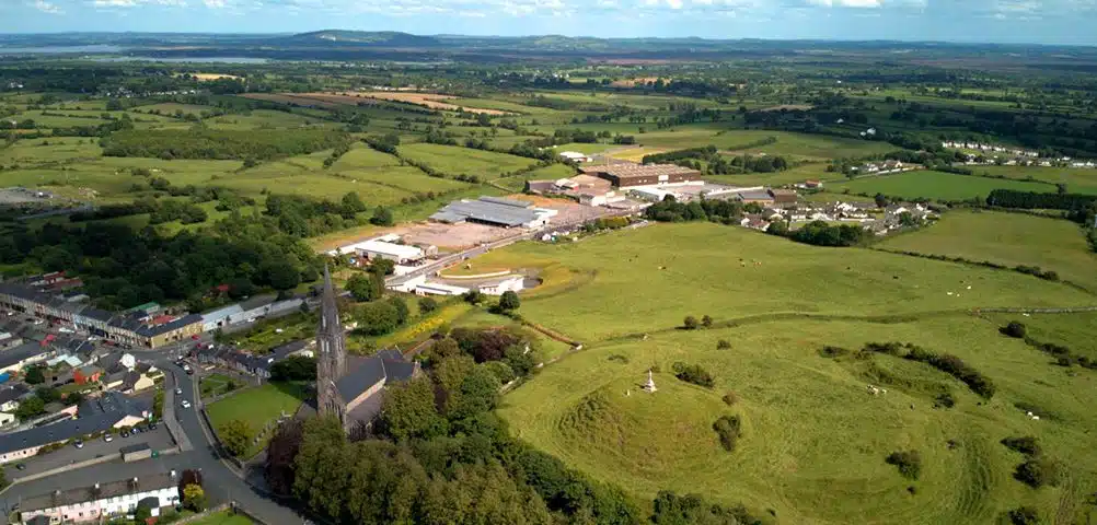 Aerial view of a rural landscape with fields, a church with a tall spire, scattered buildings, and rolling hills under a partly cloudy sky.
