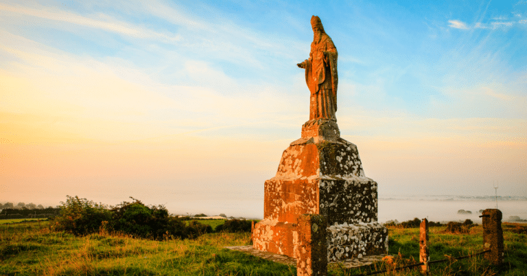 A stone statue on a tall plinth stands on a grassy hill at sunrise, overlooking fields and a misty landscape under a colourful sky.