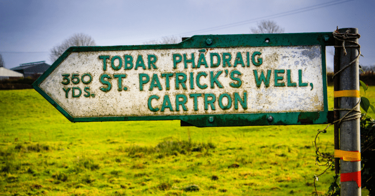 A weathered direction sign in a grassy field reads "350 yds. TOBAR PHÁDRAIG, ST. PATRICK’S WELL, CARTRON" in English and Irish.