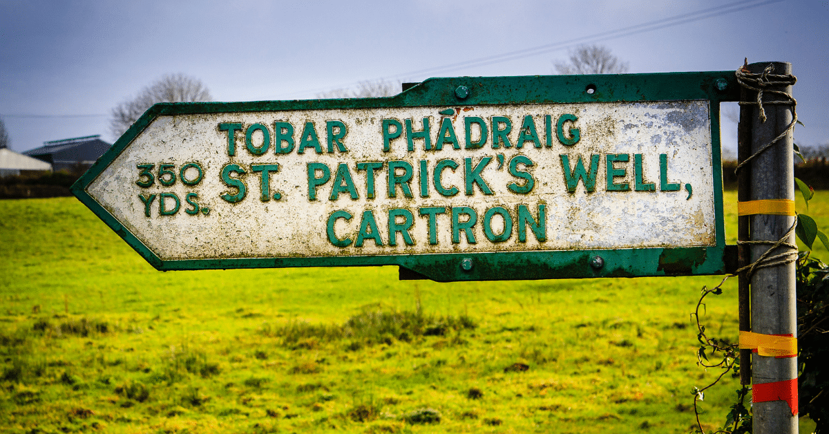 A weathered direction sign in a grassy field reads "350 yds. TOBAR PHÁDRAIG, ST. PATRICK’S WELL, CARTRON" in English and Irish.