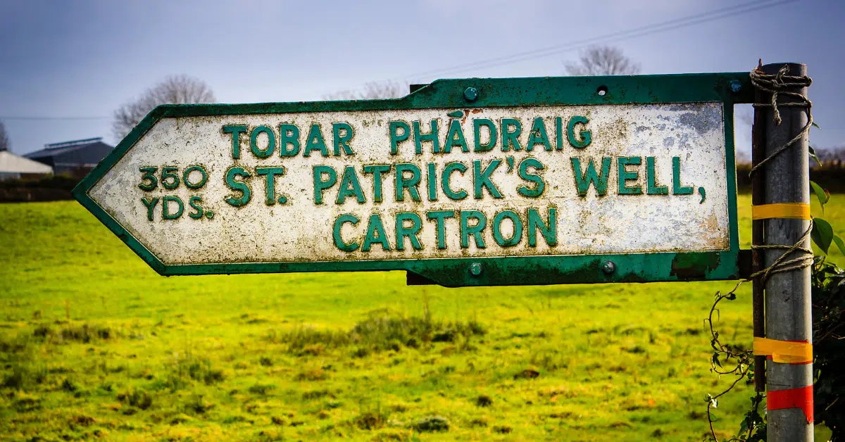 A weathered direction sign in a grassy field reads "350 yds. TOBAR PHÁDRAIG, ST. PATRICK’S WELL, CARTRON" in English and Irish.