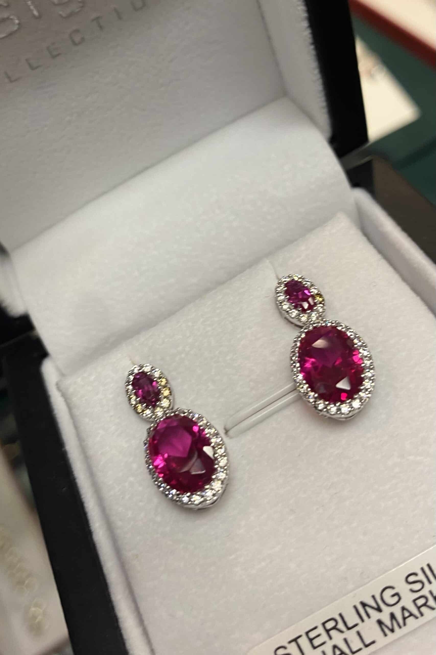 A pair of sterling silver earrings each featuring two oval-shaped pink gemstones surrounded by small clear stones, displayed in a white jewellery box.