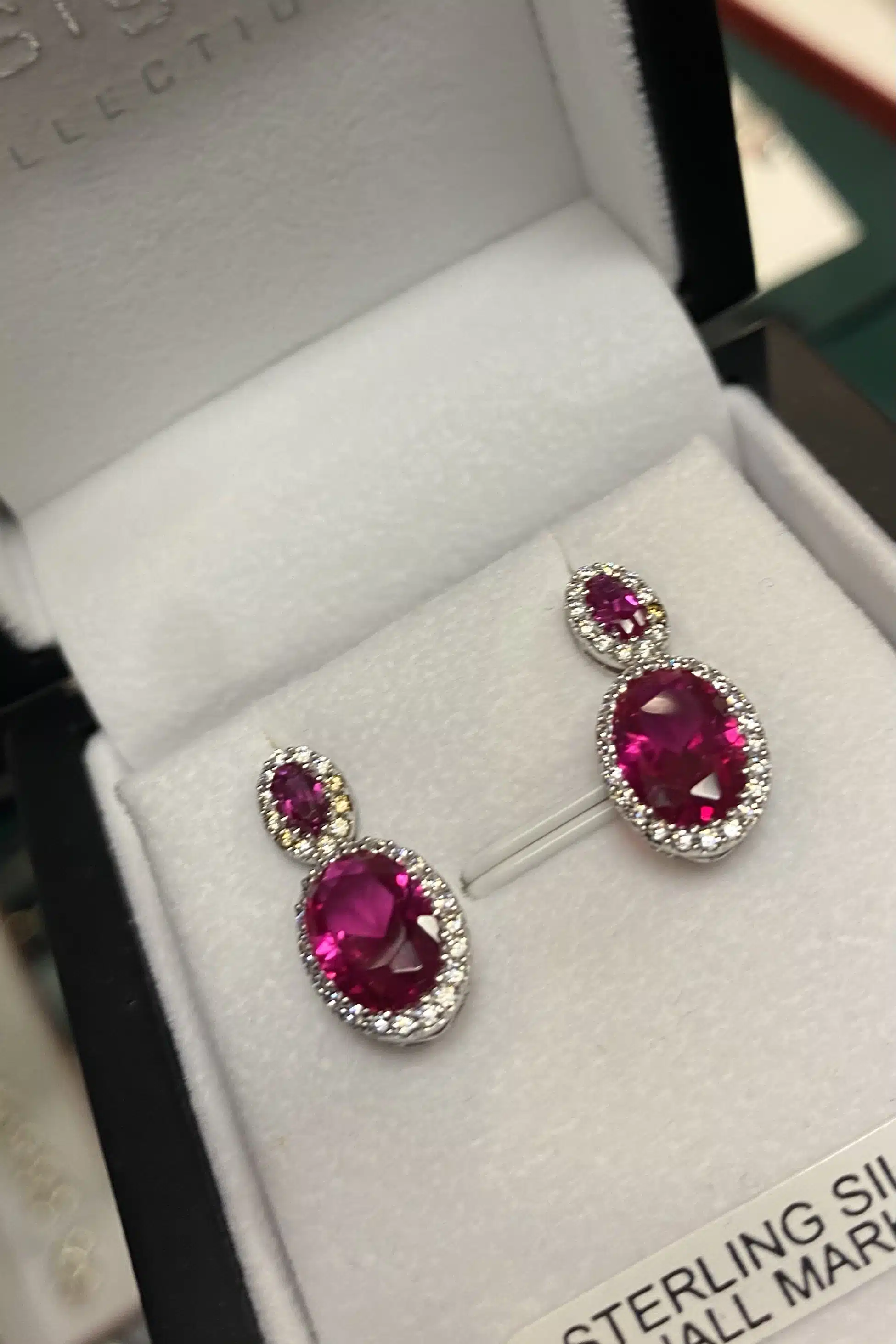 A pair of sterling silver earrings each featuring two oval-shaped pink gemstones surrounded by small clear stones, displayed in a white jewellery box.