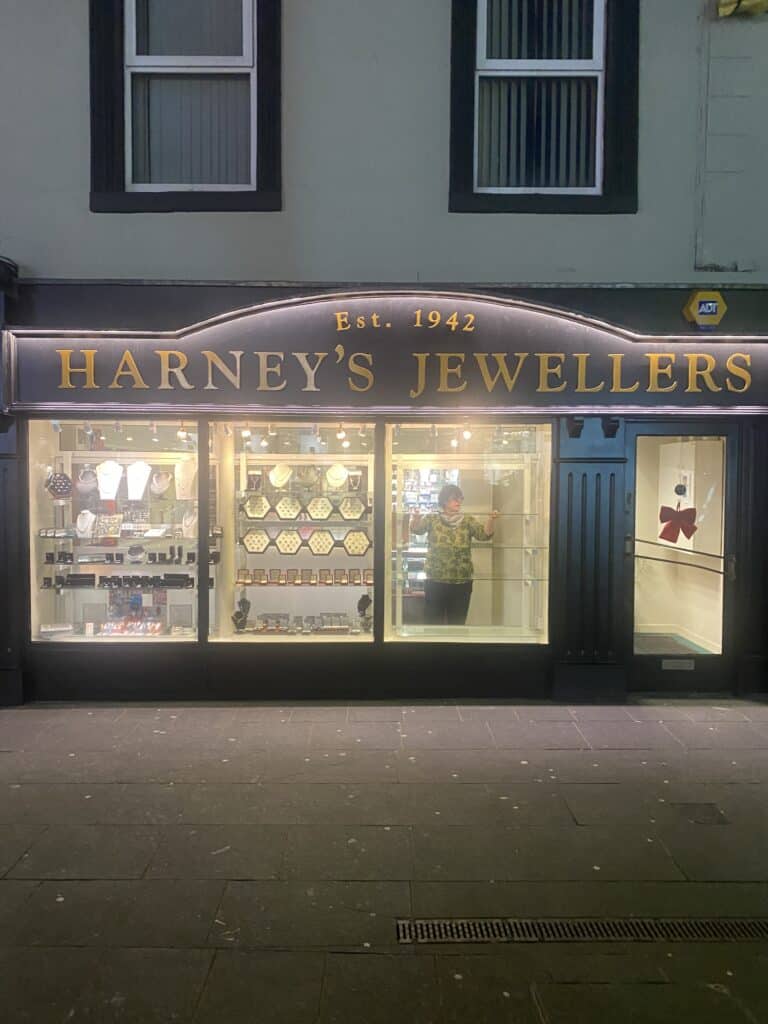 A jewellery shop called "Harney's Jewellers" with a display of necklaces and watches in the window. A person stands inside the brightly lit shop, visible through the glass. The sign says "Est. 1942.