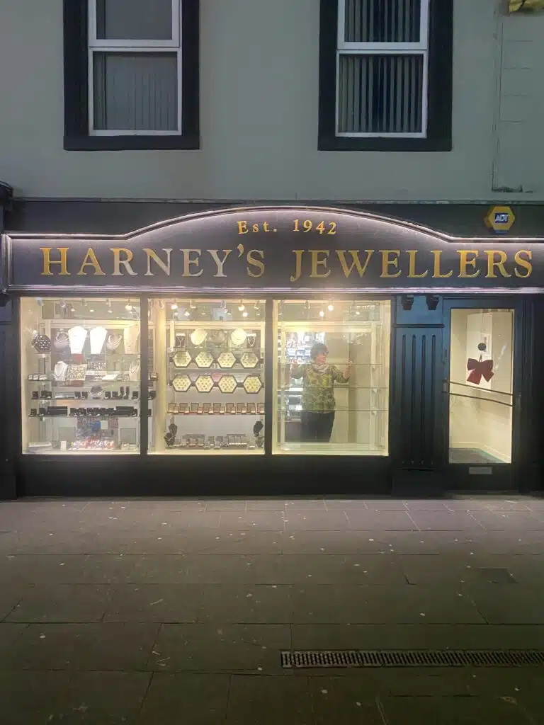 A jewellery shop called "Harney's Jewellers" with a display of necklaces and watches in the window. A person stands inside the brightly lit shop, visible through the glass. The sign says "Est. 1942.