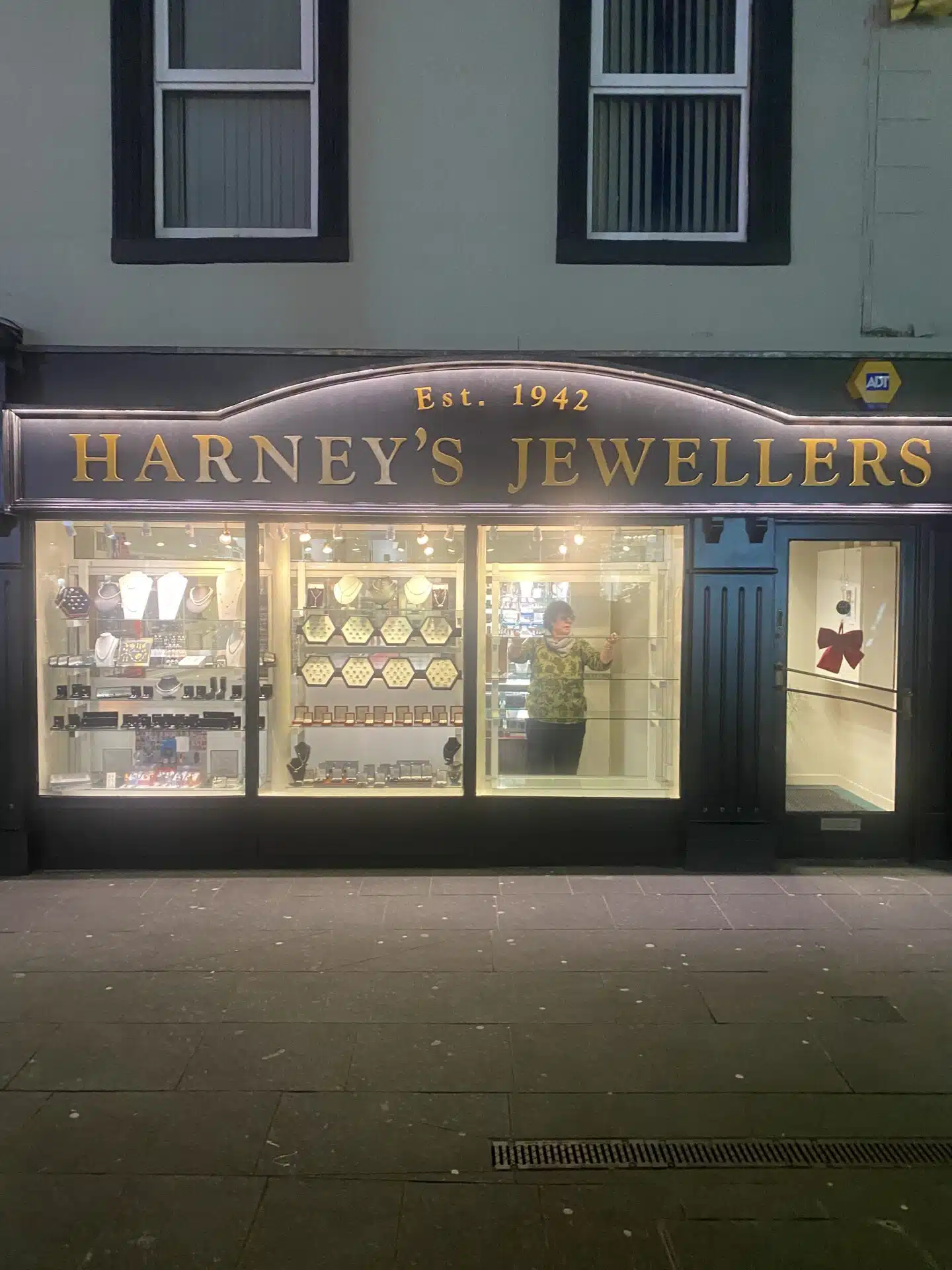 A jewellery shop called "Harney's Jewellers" with a display of necklaces and watches in the window. A person stands inside the brightly lit shop, visible through the glass. The sign says "Est. 1942.