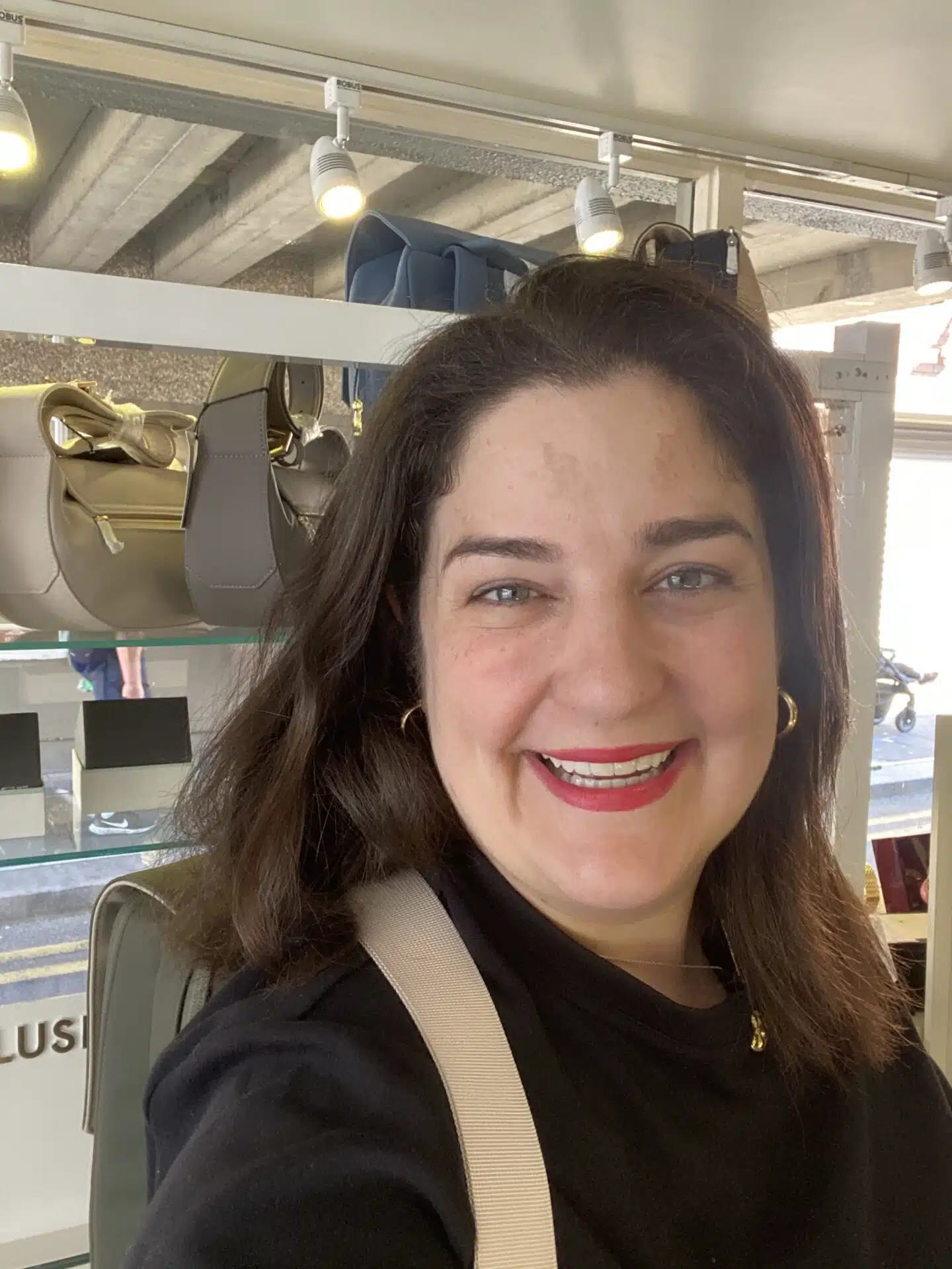 A woman with dark hair and red lipstick smiles at the camera inside a shop with handbags displayed on shelves behind her.