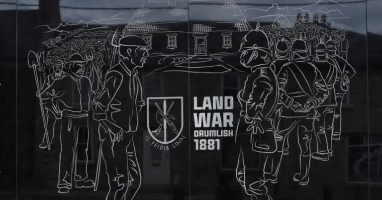 Line drawing on a plaque depicts men with tools facing armed officers in front of a building. Text reads “LAND WAR DRUMLISH 1881” with an emblem and Irish phrase, referencing an historical event.
