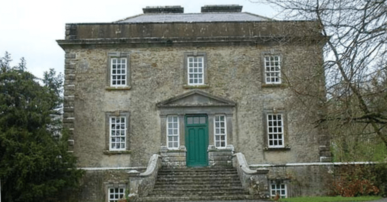 A stone Georgian-style house with a green door, tall windows, and stone steps leading to the entrance. Trees and shrubbery surround the historic building.