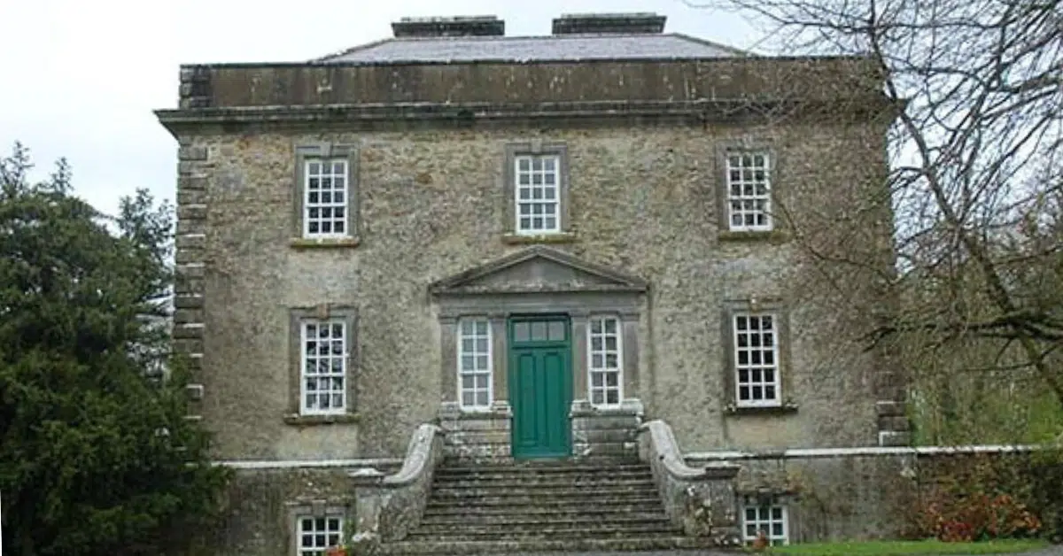 A stone Georgian-style house with a green door, tall windows, and stone steps leading to the entrance. Trees and shrubbery surround the historic building.