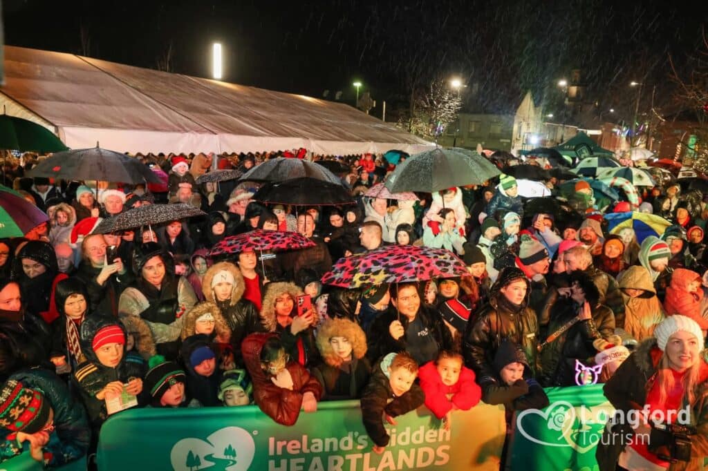 A large crowd, many holding umbrellas and wearing winter clothes, gathers at an outdoor event at night. A marquee and festive lights are visible in the background. A green banner in front reads "Ireland's Hidden Heartlands," highlighting Sustainable Tourism Longford as part of the celebration.