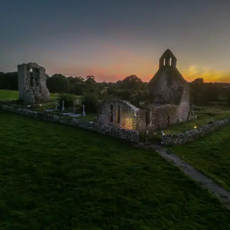 An aerial view of ancient stone ruins and grave markers surrounded by a stone wall, set against a grassy landscape at sunset with a colourful sky in the background.