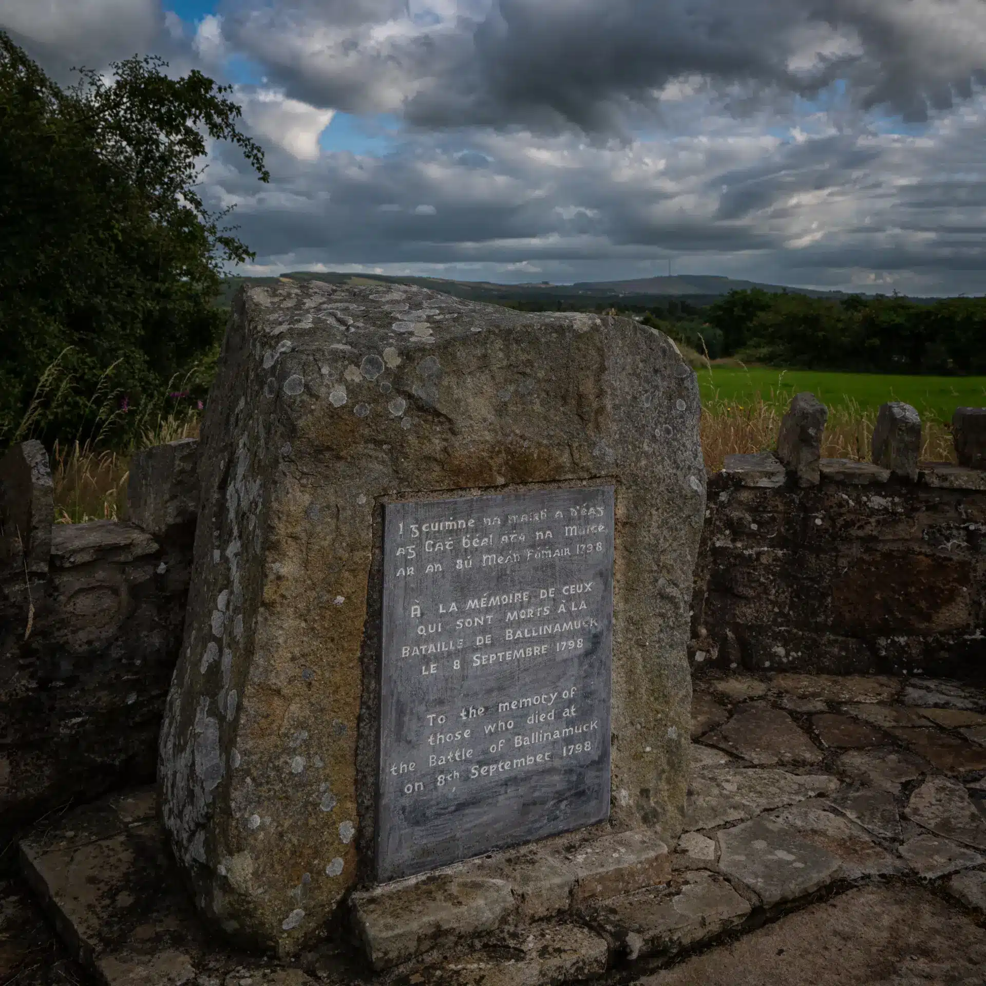 A stone memorial with an inscribed plaque stands on a stone platform, surrounded by a low stone wall, overlooking green fields and hills under a cloudy sky.