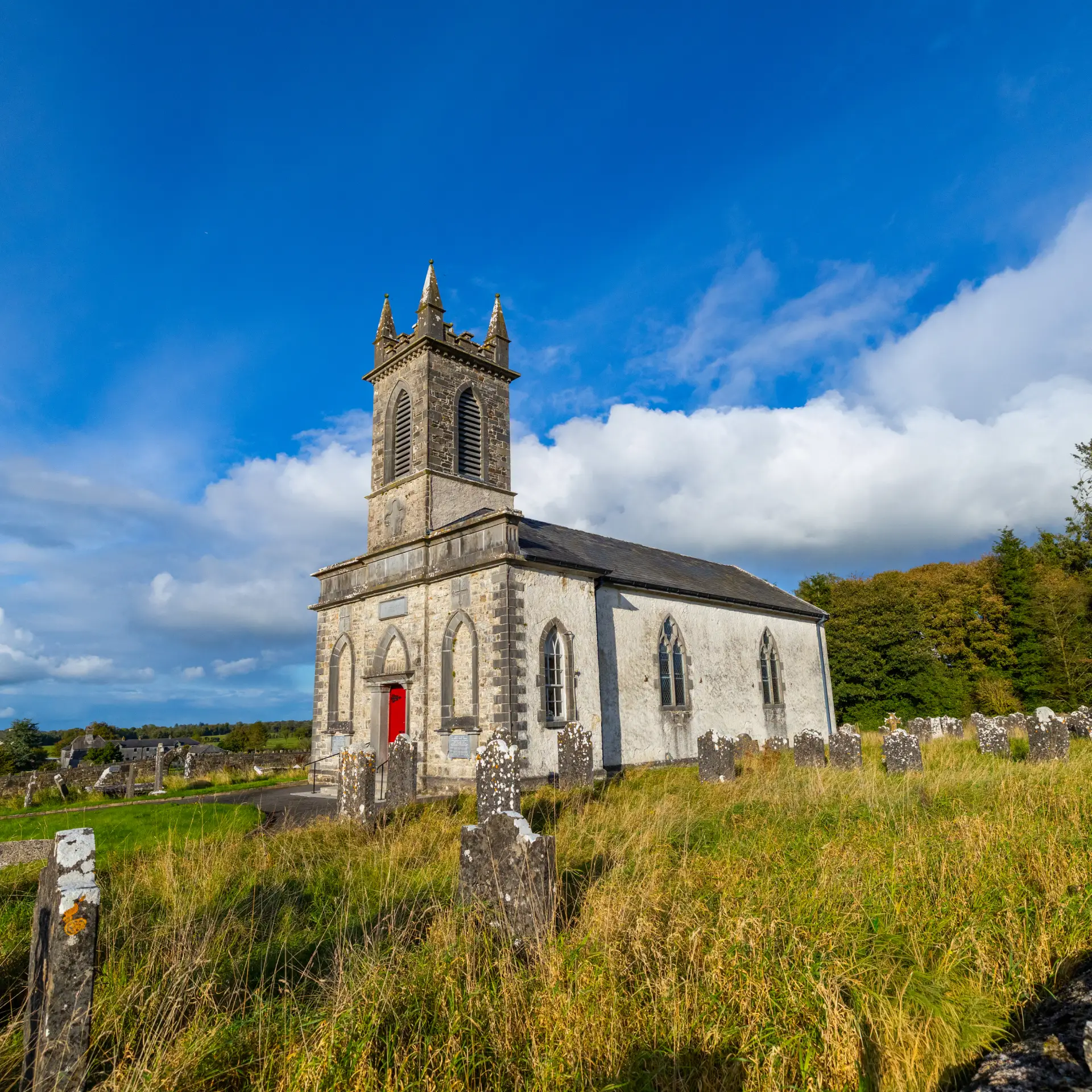 A stone church with a tall clock tower and a bright red door stands surrounded by old gravestones and overgrown grass, under a blue sky with scattered clouds. Trees are visible in the background.