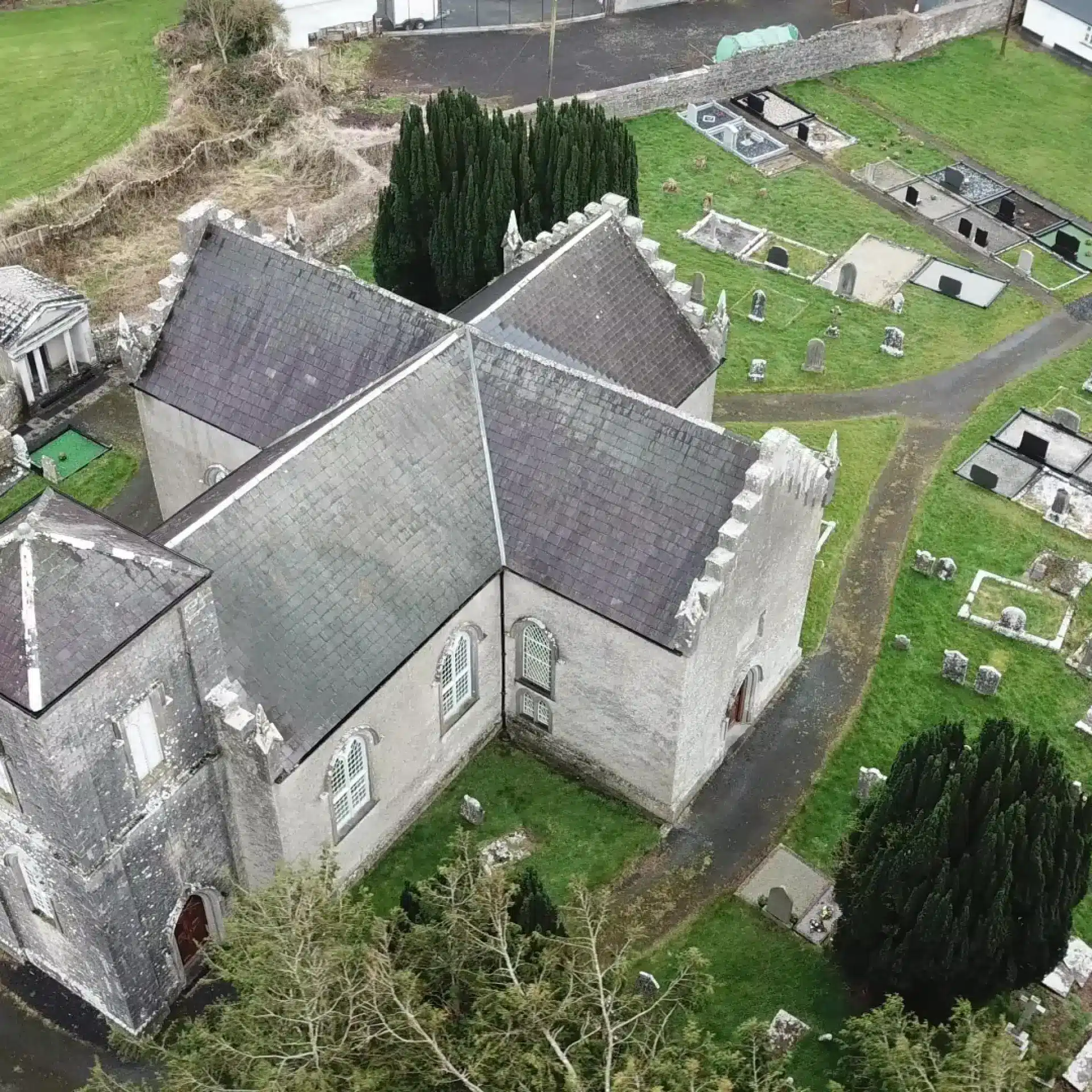 Aerial view of an old stone church with a slate roof, surrounded by a graveyard with headstones and trees, set in a green landscape with paths and nearby buildings.