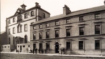 Black-and-white photo of a large, historic stone building with a clock, several windows, and people standing on the pavement out front.