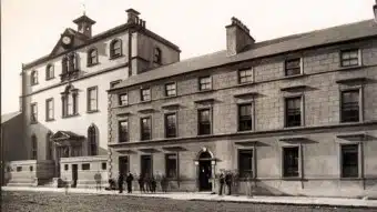 Black-and-white photo of a large, historic stone building with a clock, several windows, and people standing on the pavement out front.