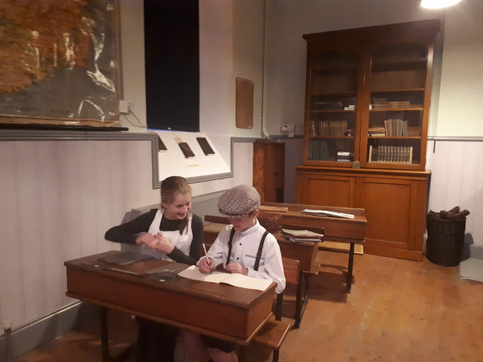 Two children in vintage clothing sit at wooden desks in an old-fashioned classroom, smiling and writing. A map, bookcase, and old books are visible in the background, evoking a historical school setting.