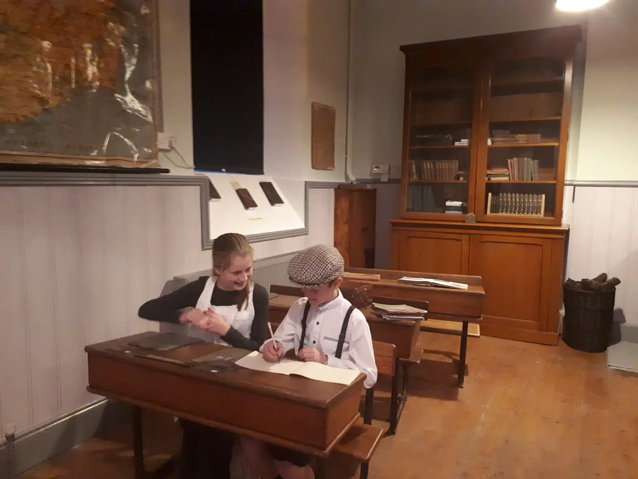 Two children in vintage clothing sit at wooden desks in an old-fashioned classroom, smiling and writing. A map, bookcase, and old books are visible in the background, evoking a historical school setting.