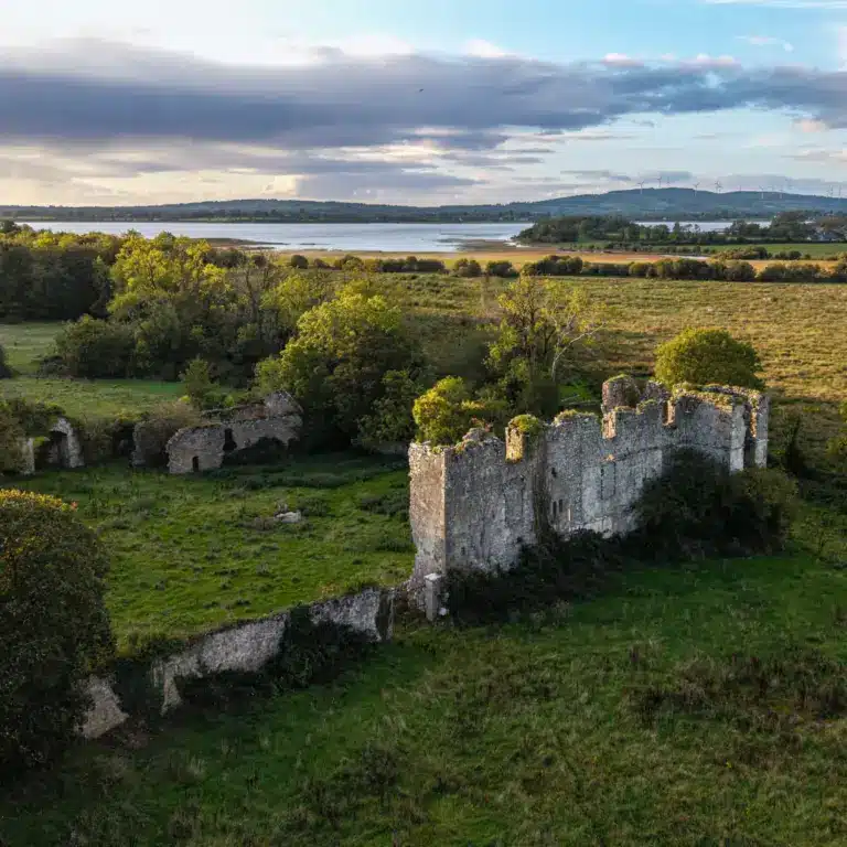 Aerial view of an old stone castle ruin surrounded by green fields and trees, with a lake and rolling hills in the background under a partly cloudy sky.