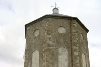 An old, weathered octagonal stone tower with boarded-up windows and a small roof topped by a weathercock, set against a cloudy sky.