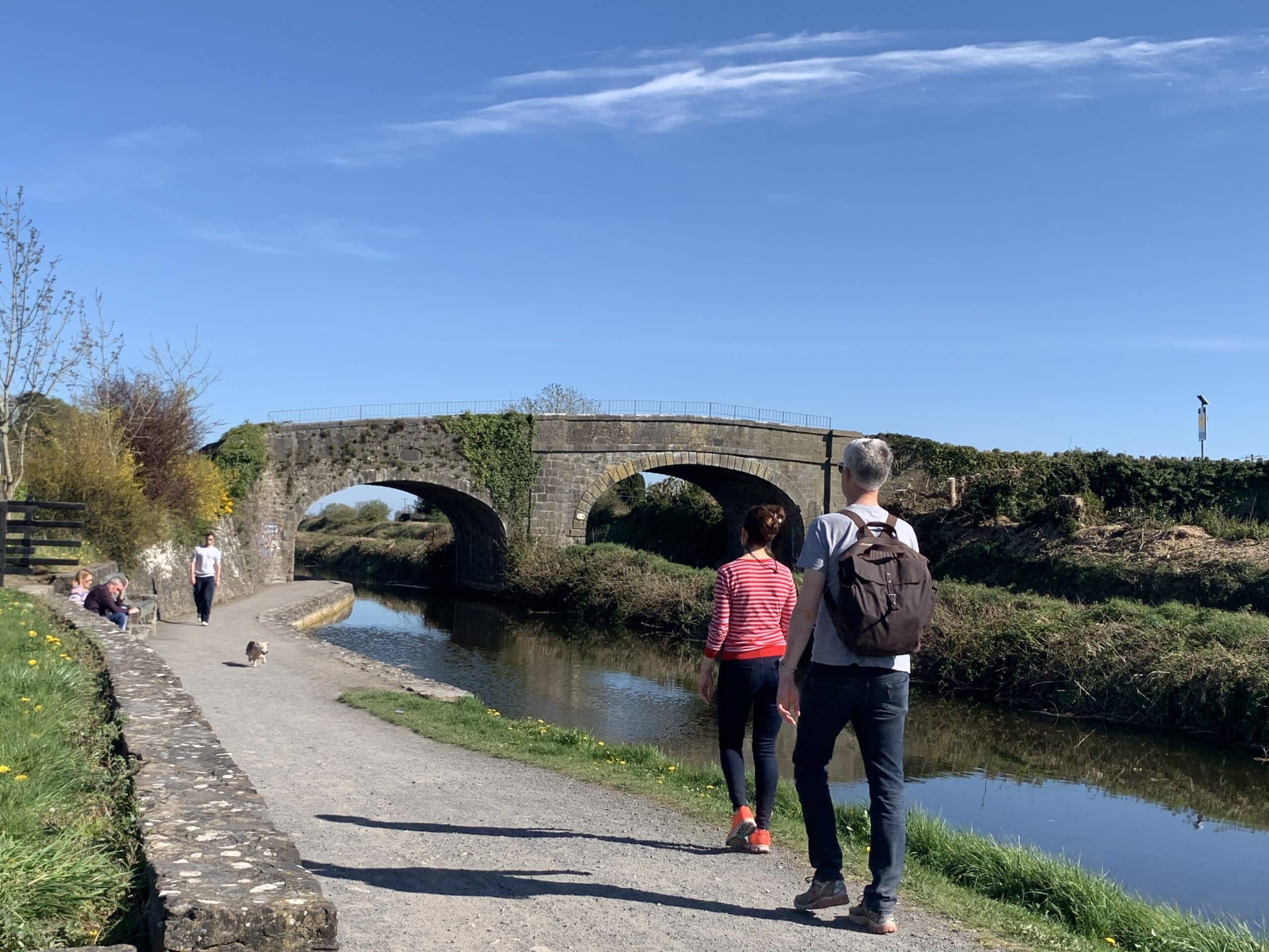 People walk along a canal towpath under a clear blue sky, passing a stone bridge. Some are accompanied by a dog, with grass and trees lining the water on both sides.