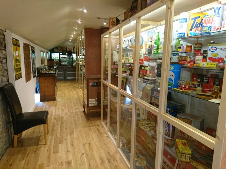 A museum corridor with wooden floors and vintage food packaging displayed behind glass cases. A black chair sits along the left wall near various posters.