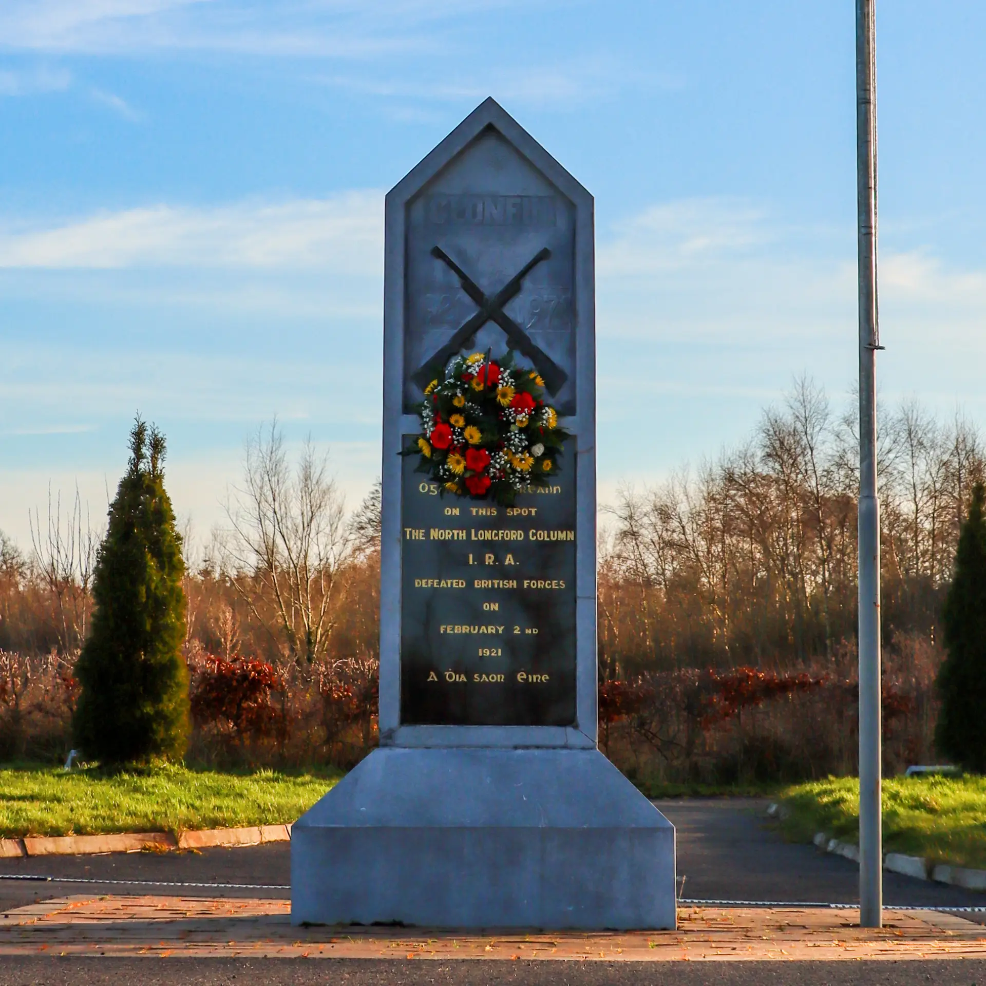 A stone memorial obelisk with a wreath of flowers and engraved text stands in an open area, surrounded by trees and shrubbery on a sunny day.