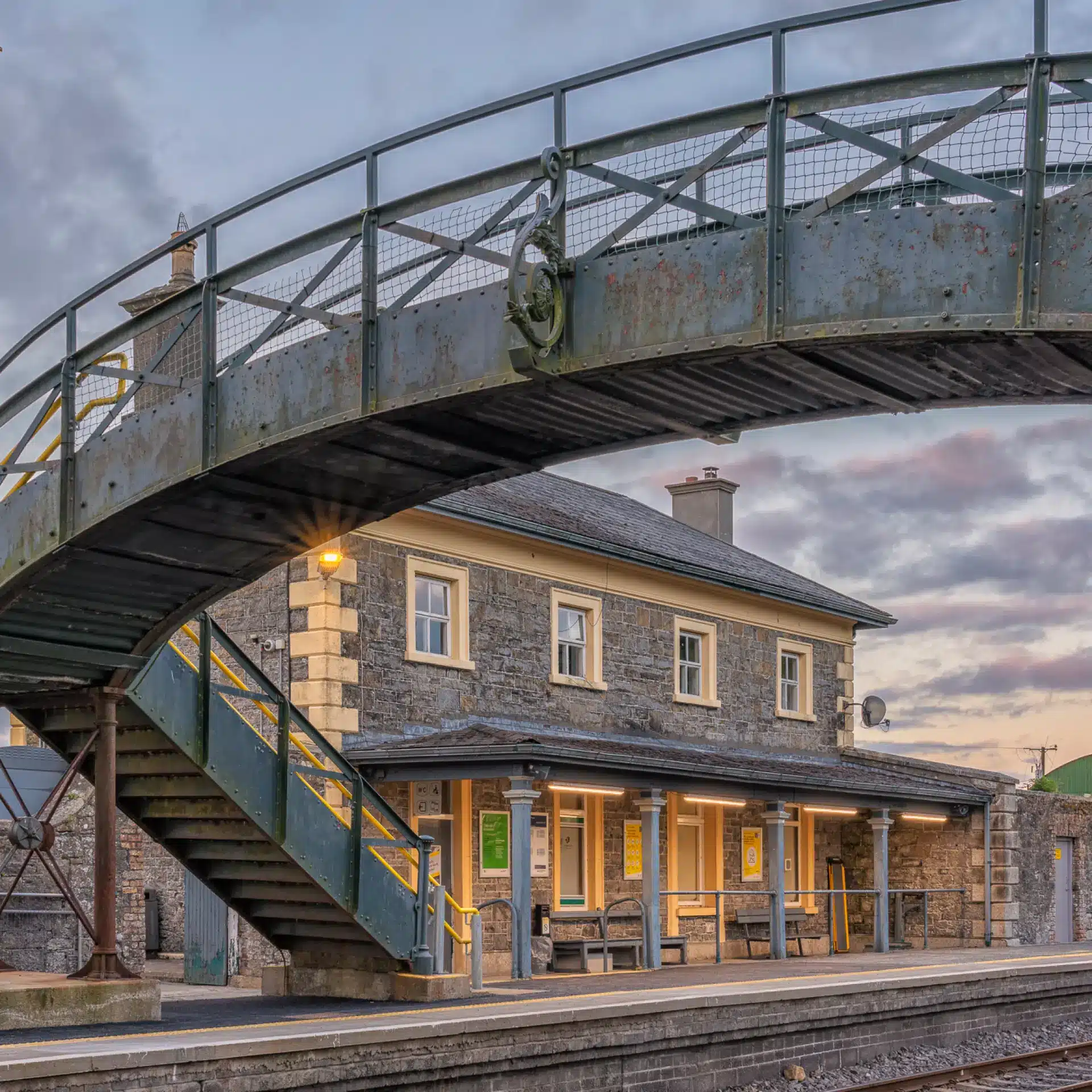 A stone railway station with yellow-trimmed doors and windows sits beneath an old, green metal footbridge. The sky is cloudy with a hint of sunset, and the platform is empty.
