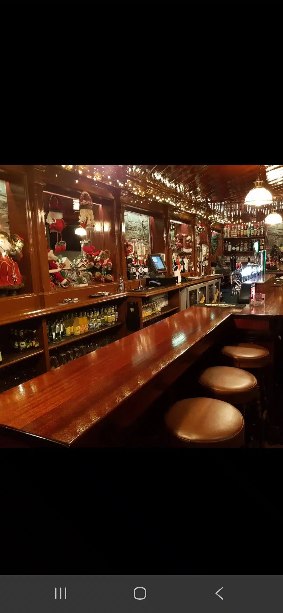 A polished wooden bar with three stools, festive Christmas decorations, fairy lights, and shelves of glasses and bottles. The bar area is warmly lit, creating a cosy atmosphere.