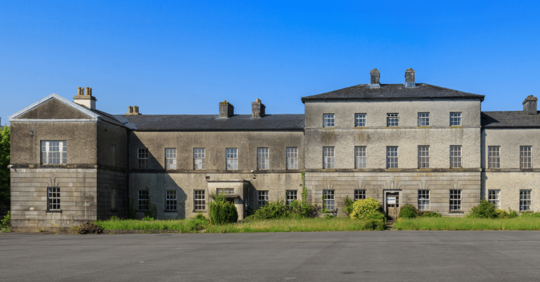 A large, old, grey stone building with many windows and chimneys, surrounded by overgrown grass and shrubs under a clear blue sky.
