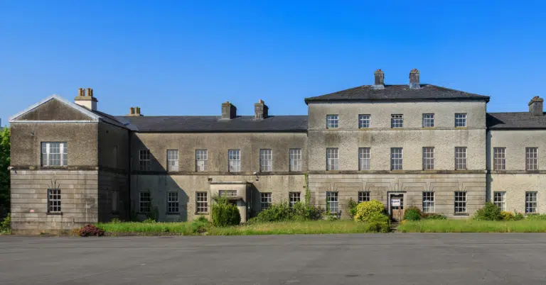 A large, old, grey stone building with many windows and chimneys, surrounded by overgrown grass and shrubs under a clear blue sky.