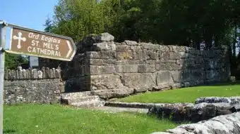 A weathered stone wall stands on grass near a wooden sign pointing to St. Mel's Cathedral, with trees in the background and sunlight illuminating the scene.
