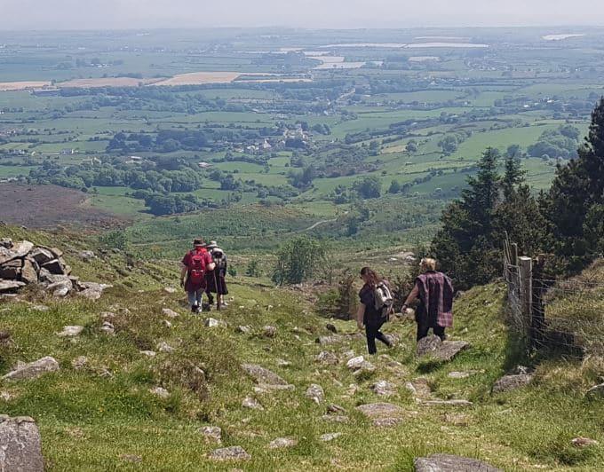 Four people hike up a grassy, rocky hill with a scenic, expansive valley and farmland visible in the background under a partly cloudy sky.