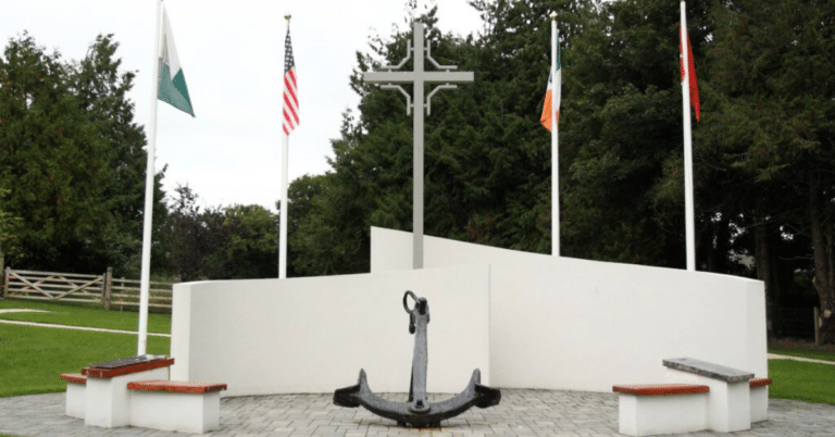 A memorial with a large anchor, three benches, a tall cross, and four flagpoles displaying different national flags, set in a landscaped park area with trees and grass in the background.
