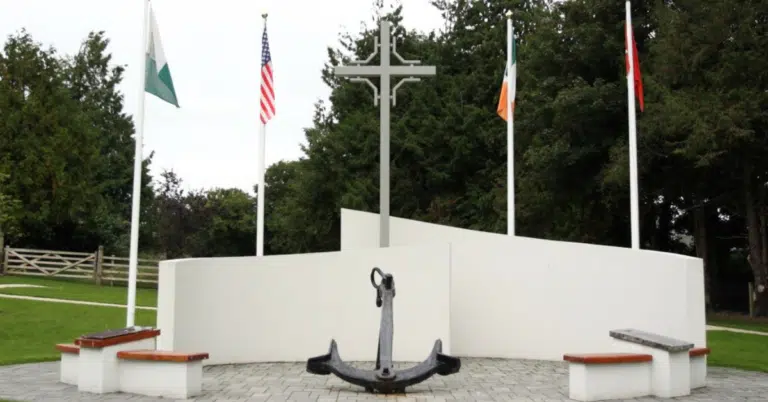 A memorial with a large anchor, three benches, a tall cross, and four flagpoles displaying different national flags, set in a landscaped park area with trees and grass in the background.