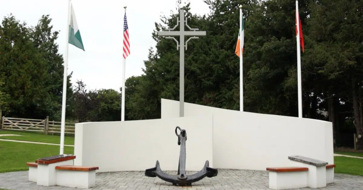 A memorial with a large anchor, three benches, a tall cross, and four flagpoles displaying different national flags, set in a landscaped park area with trees and grass in the background.