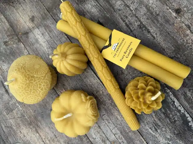Several handmade yellow beeswax candles of various shapes, including tapers, a pine cone, a pumpkin, a rose, and a round floral design, are arranged on a rustic wooden surface.