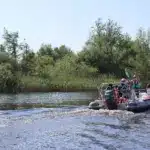 A small inflatable motorboat with several people on board moves along a calm river, leaving ripples in the water—an ideal setting for Family Activities Longford. Lush green trees and reeds line the riverbank under a clear blue sky.