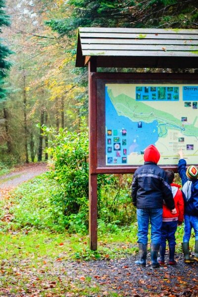 Four children and one adult, all in jackets and boots, stand in front of a large map sign in a forest, checking the trail information. Autumn leaves cover the ground and trees line the path beside them.