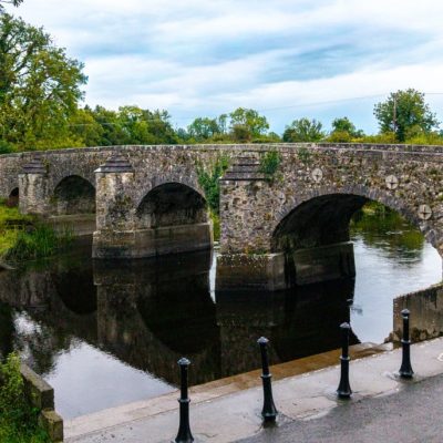 A stone arch bridge crosses a calm river surrounded by green trees. In the foreground, a roadside sign and black bollards line the edge of the road leading towards the bridge. The sky is cloudy.
